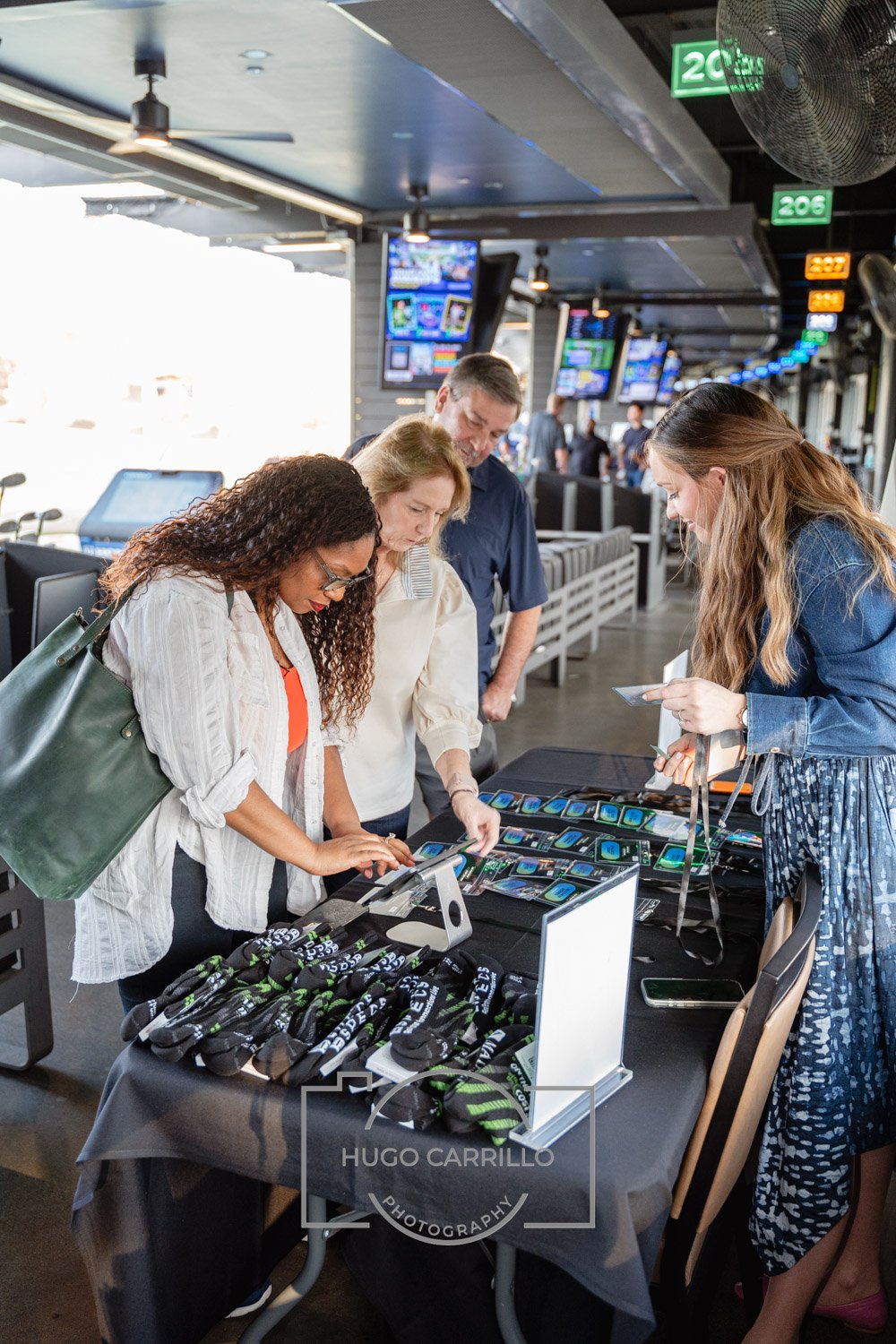 People at a merchandise table with promotional items, at an airport or indoor location with multiple TV screens and seating area.