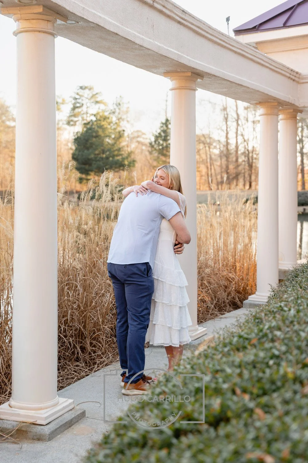 A woman in a white dress hugging a man in a light blue shirt under a white columned pavilion in a park with tall grass and trees in the background during sunset.