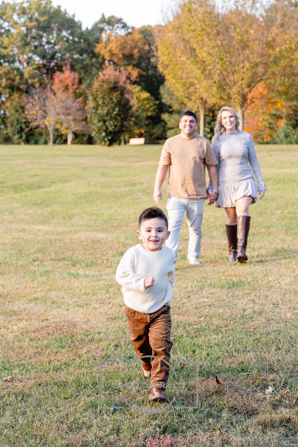 A young boy in a white sweater and brown pants is running towards the camera in a grassy park, with a man and woman holding hands and walking behind him, surrounded by trees with autumn foliage.