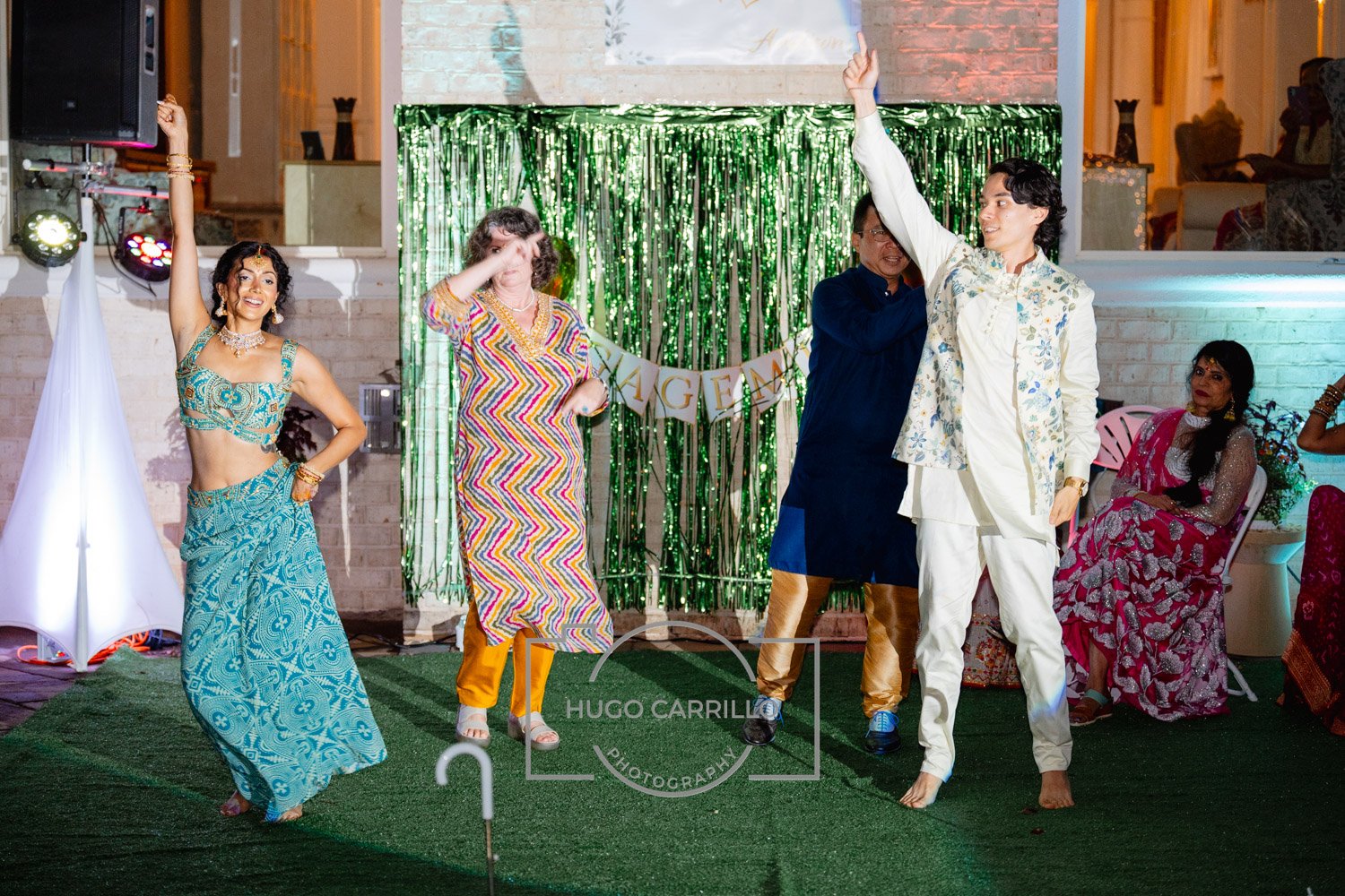 People dancing and celebrating at a gathering with colorful outfits and decorations, including a green backdrop and a banner that says 'ENGAGEMENT'.