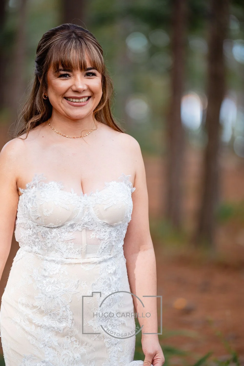 Smiling woman in a white lace strapless wedding dress standing outdoors in a forested area.
