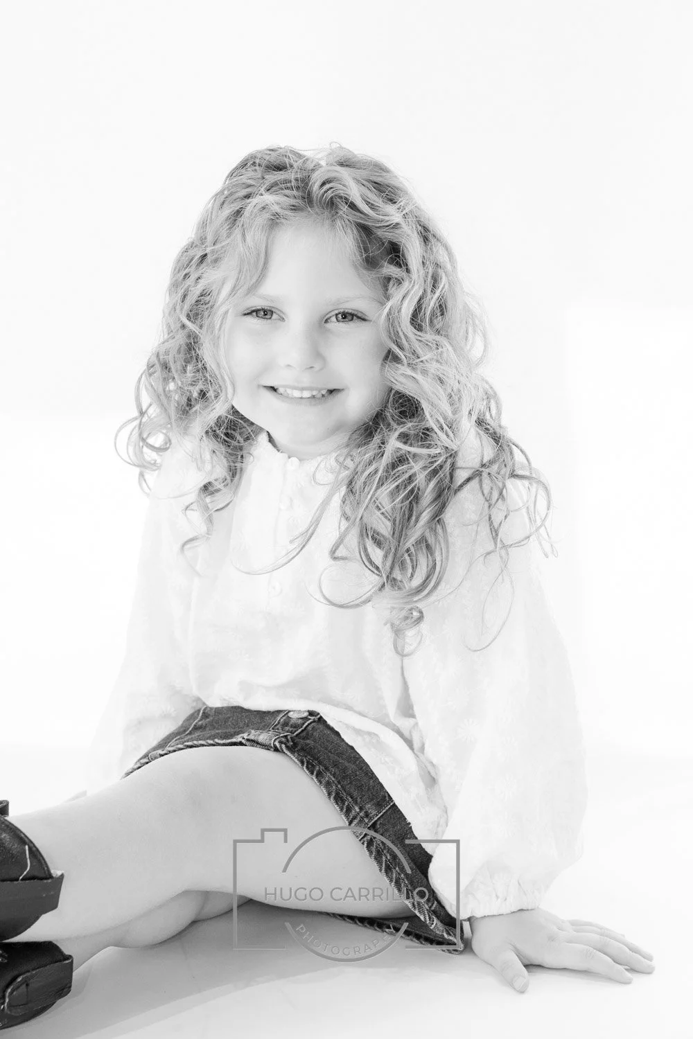 A young girl with curly hair smiling at the camera, sitting with her legs crossed, wearing a light-colored blouse and denim shorts, against a plain background.