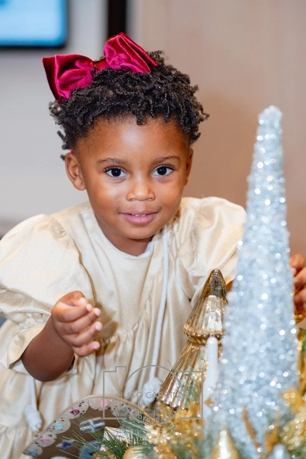 A young girl with curly hair wearing a cream-colored dress and a large red velvet bow in her hair, looking at the camera with a slight smile, surrounded by holiday decorations including a silver glittery cone and other festive ornaments.