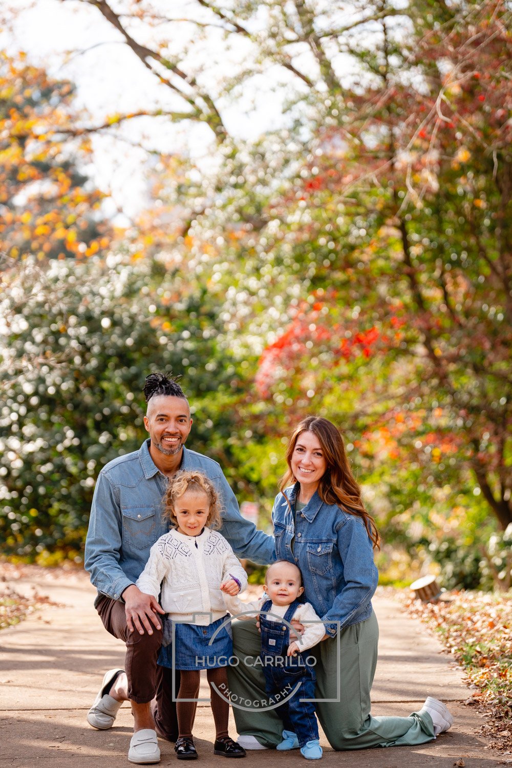 A family of four, two parents and two young children, smiling and posing outdoors on a walk in a park during autumn, with colorful fall leaves on trees.