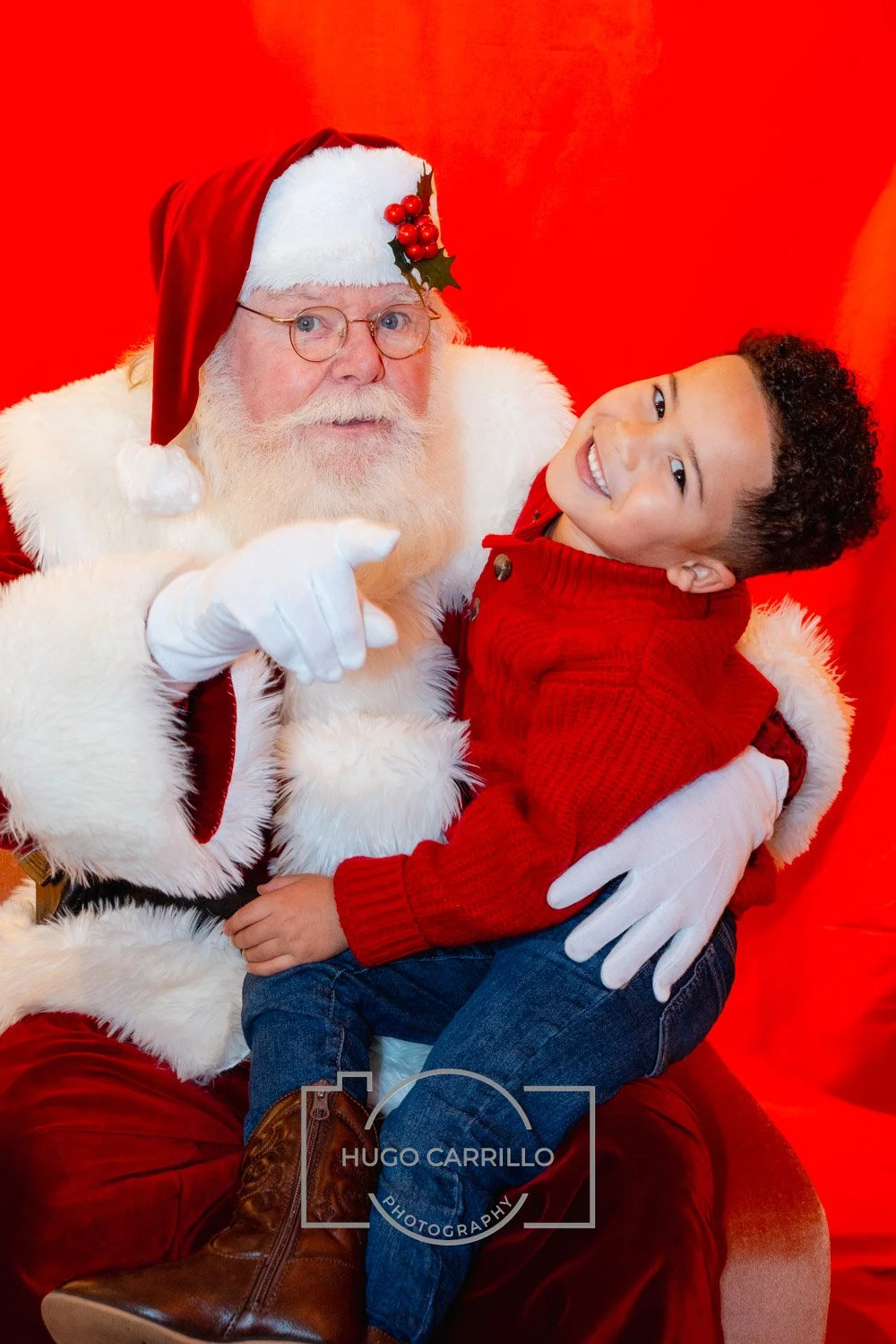 Santa Claus with glasses, white beard, red suit, and a holly decoration on his hat, sitting with a young boy in a red sweater, both smiling at the camera against a red background.