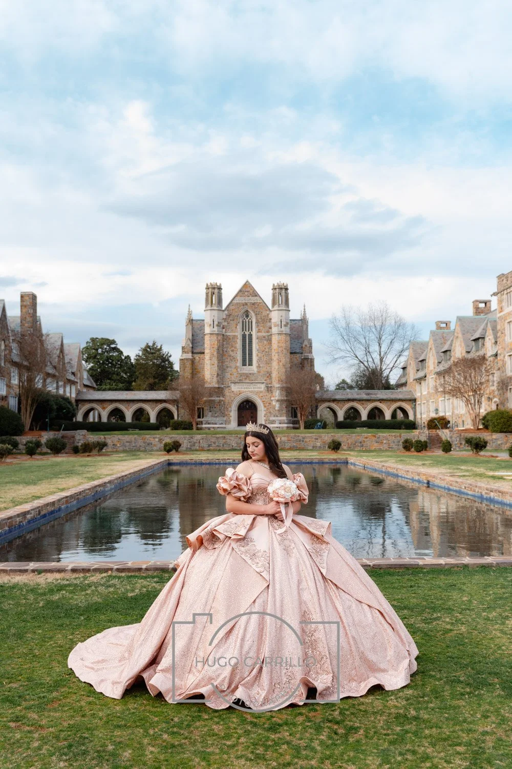 A quinceañera in an elegant pink ball gown and tiara standing on a lawn in front of a pond with a large historic building resembling a castle or cathedral in the background.