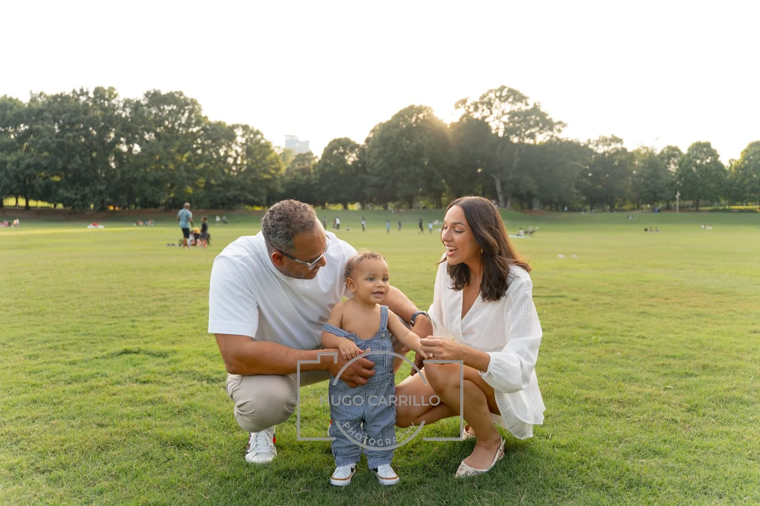 Family enjoying time together in a park with green grass and trees in the background during sunset, with adults and children sitting and walking.