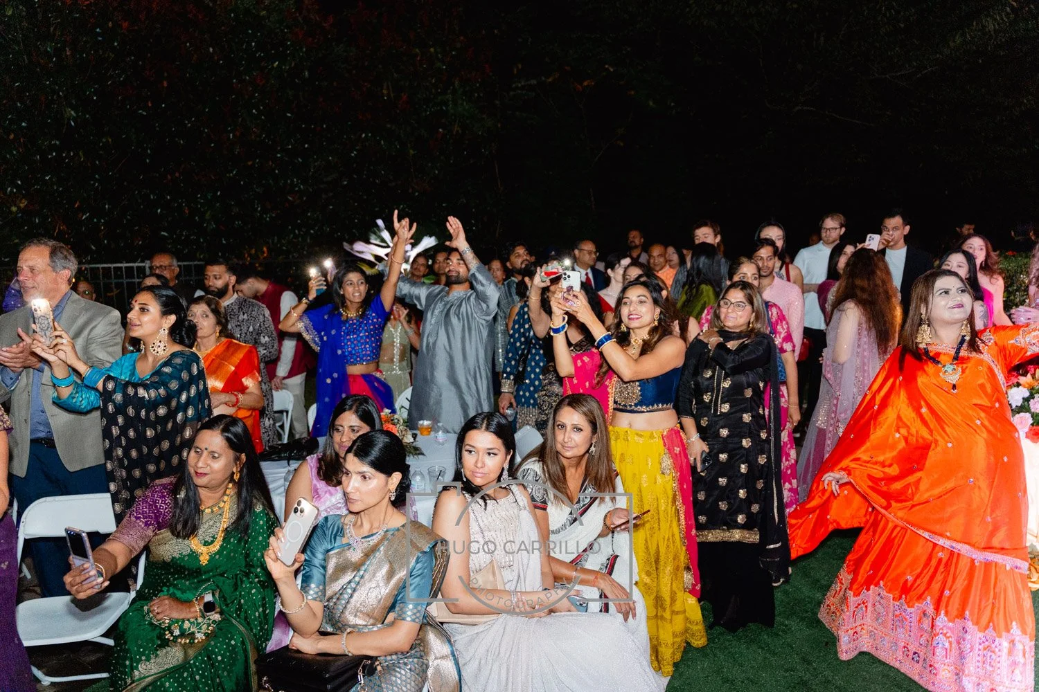 A large group of people at an outdoor festive gathering at night, many wearing colorful traditional Indian attire, some taking photos or videos, with a woman in an orange dress posing in the front right.