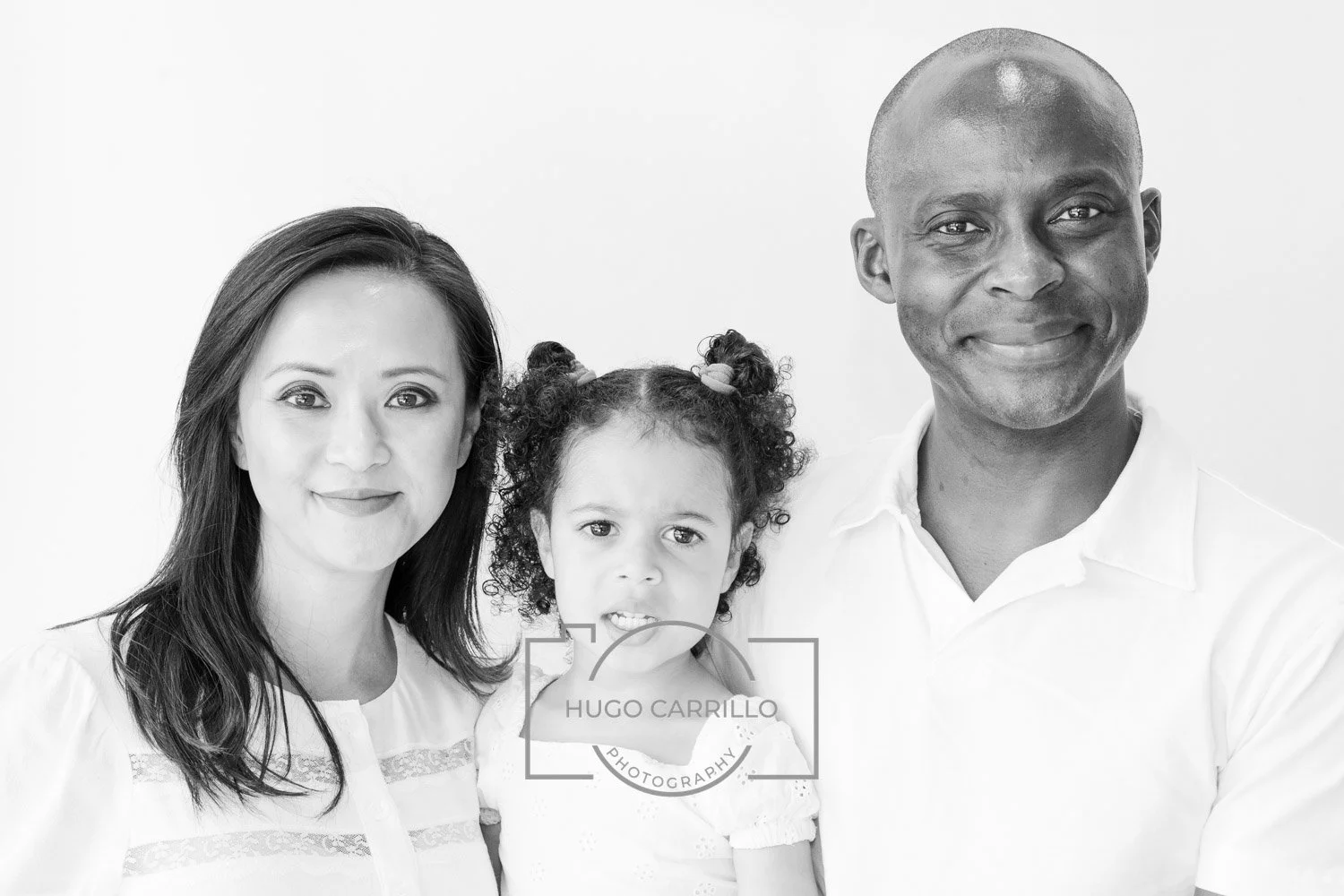 Black and white photo of three people: a woman with dark hair, a young girl with curly hair and pigtails, and a man with a shaved head, all smiling and posing together.