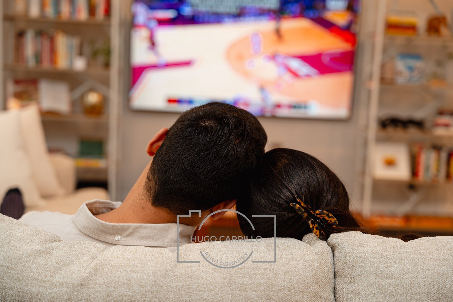 A couple sitting close together on a sofa watching a basketball game on television, with their heads resting on each other's shoulders.