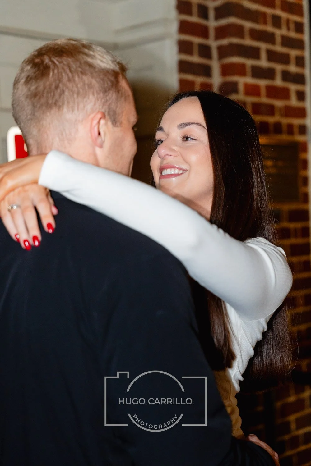 A woman and a man dance closely indoors with a brick wall and a wooden wall in the background.