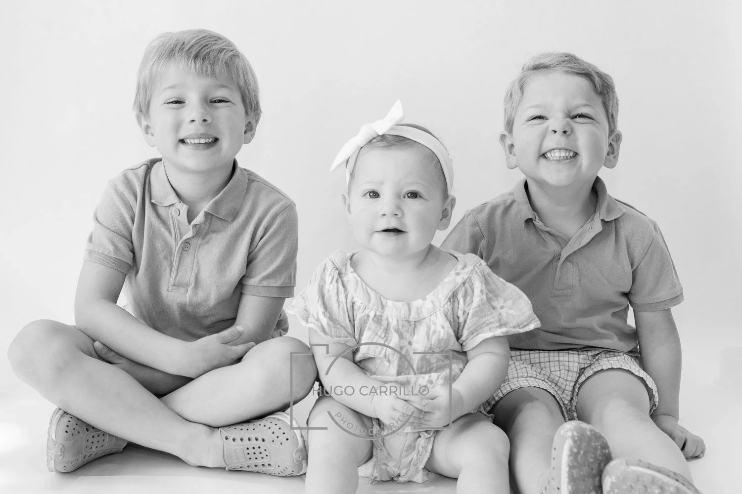 Three cheerful children, two boys and one girl, sitting together against a plain background, smiling and showing teeth.