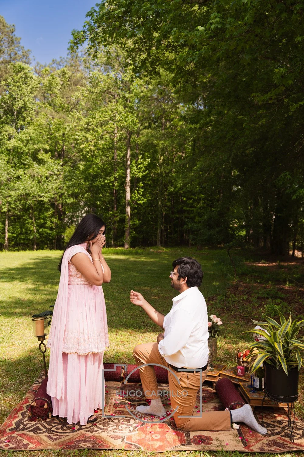 A man proposing marriage to a woman outdoors in a garden, with trees and greenery in the background. The woman is standing, covering her mouth in surprise, while the man is kneeling on one knee with a ring in his hand. They are on a decorative rug wi