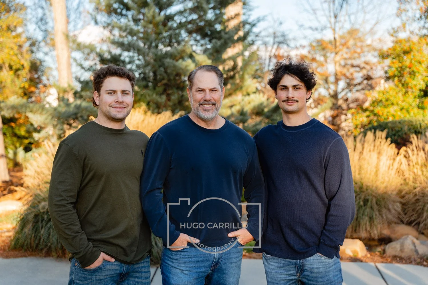 Three men standing outdoors in autumn, smiling, with trees and shrubs in the background.