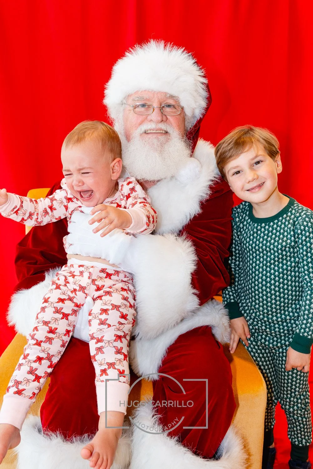 A young girl crying on Santa Claus's lap, with an older boy standing next to them, all against a red backdrop, during a Christmas event.