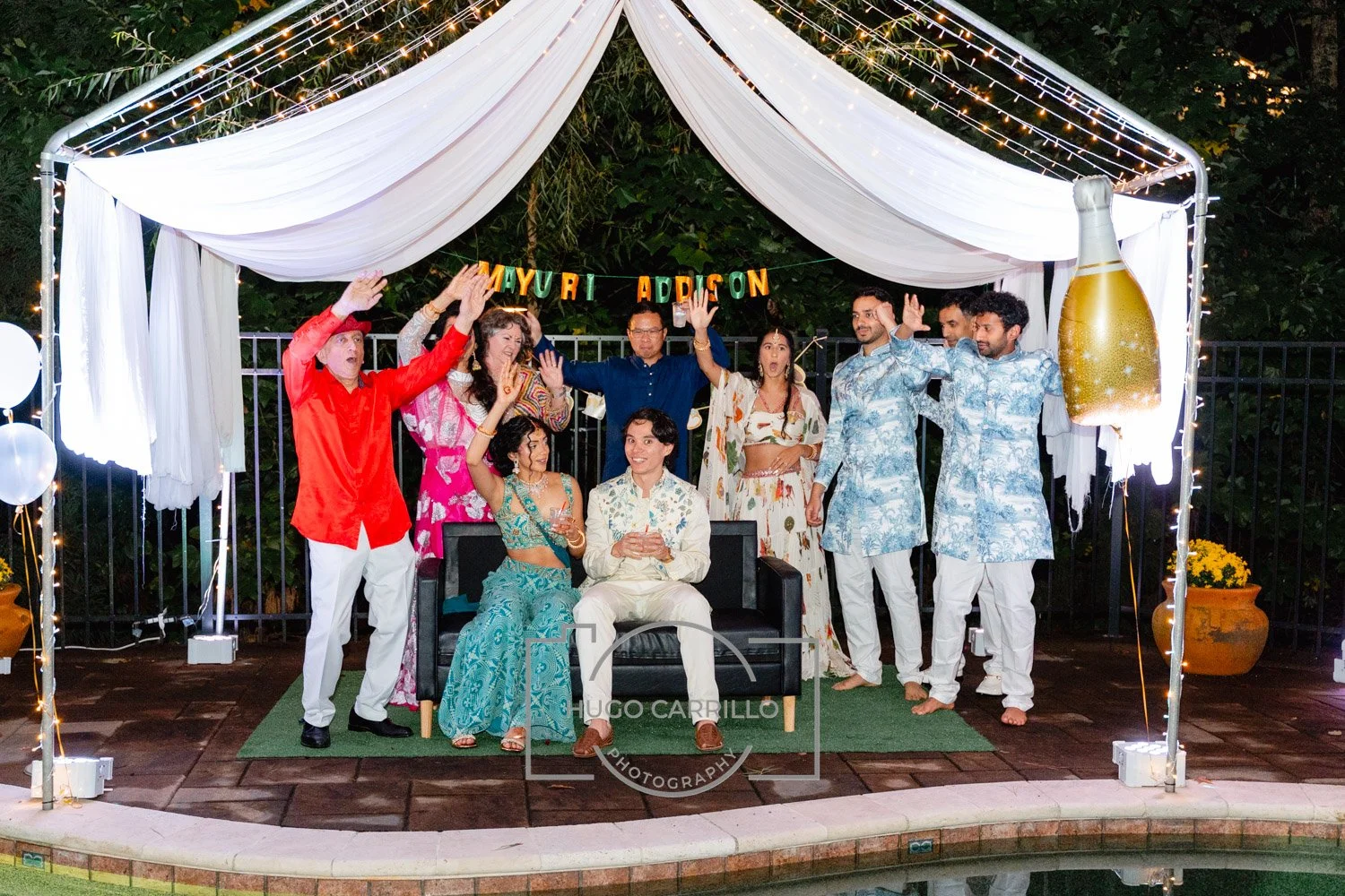 A group of people celebrating outdoors under a decorated canopy with string lights and white drapes, with a large champagne bottle decoration on the right, and a banner reading 'Mayuri Addition' above. Some are standing, some sitting on a sofa, weari