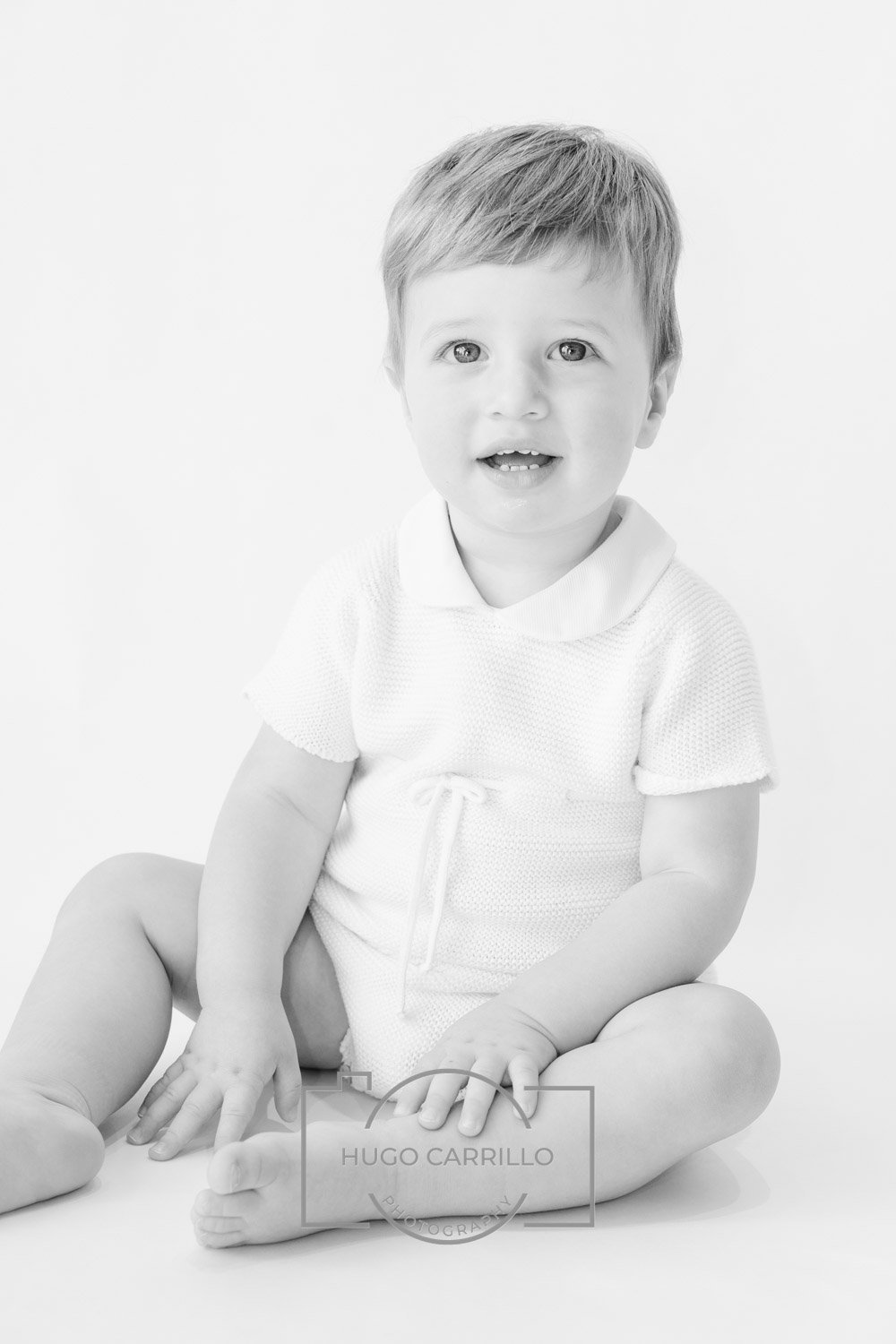 A young child sitting barefoot on the floor, wearing a light-colored, short-sleeved outfit, with a surprised or confused expression, in a studio photo with a plain white background.