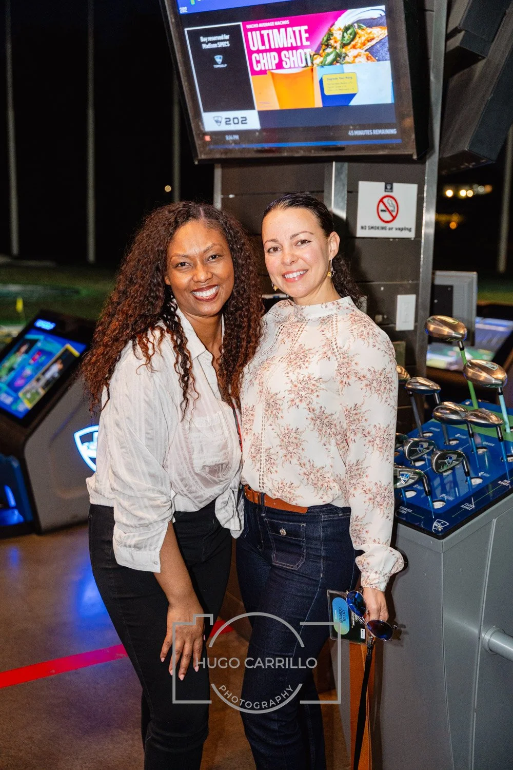Two women standing inside a golf entertainment center, smiling for the camera. There are golf clubs and screens in the background.