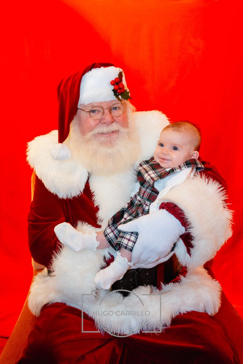 A man dressed as Santa Claus holding a smiling baby against a red background.