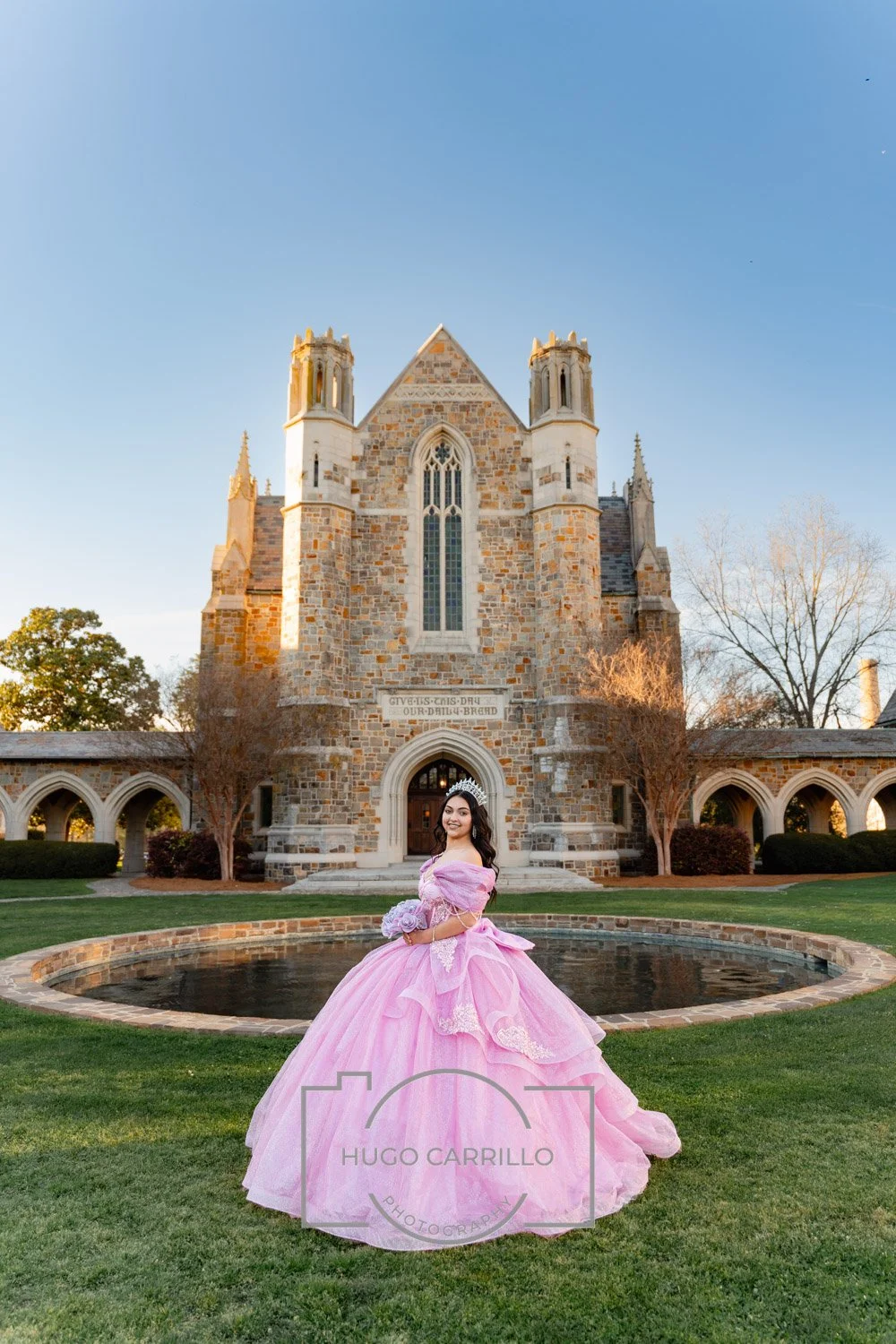 A quinceañera in a pink gown and tiara standing in front of a stone castle with a small pond and well-manicured lawn.