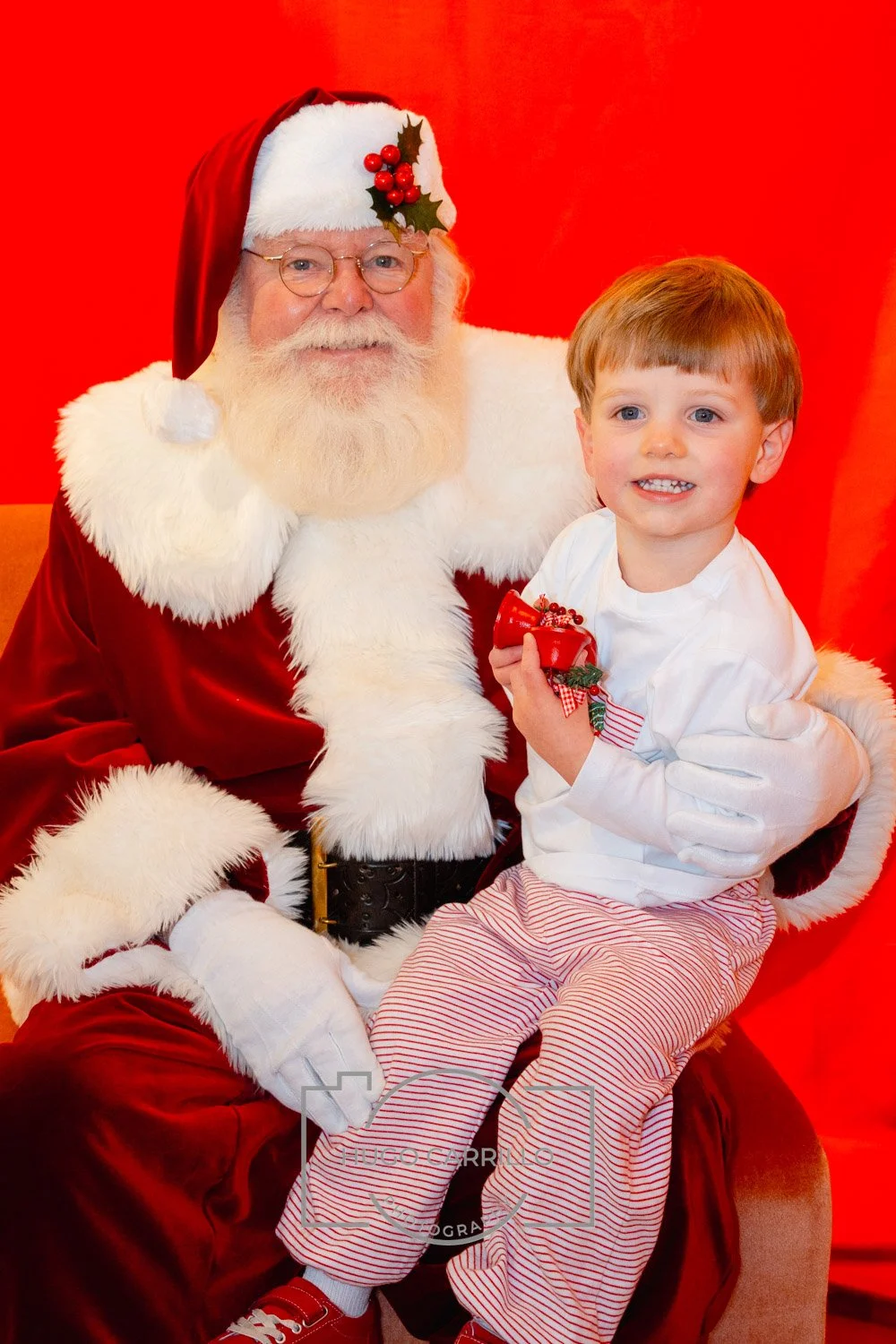 A young boy sitting on Santa Claus's lap holding a small red gift, with Santa wearing a red suit, white beard, glasses, and a Santa hat with a holly decoration, against a red background.