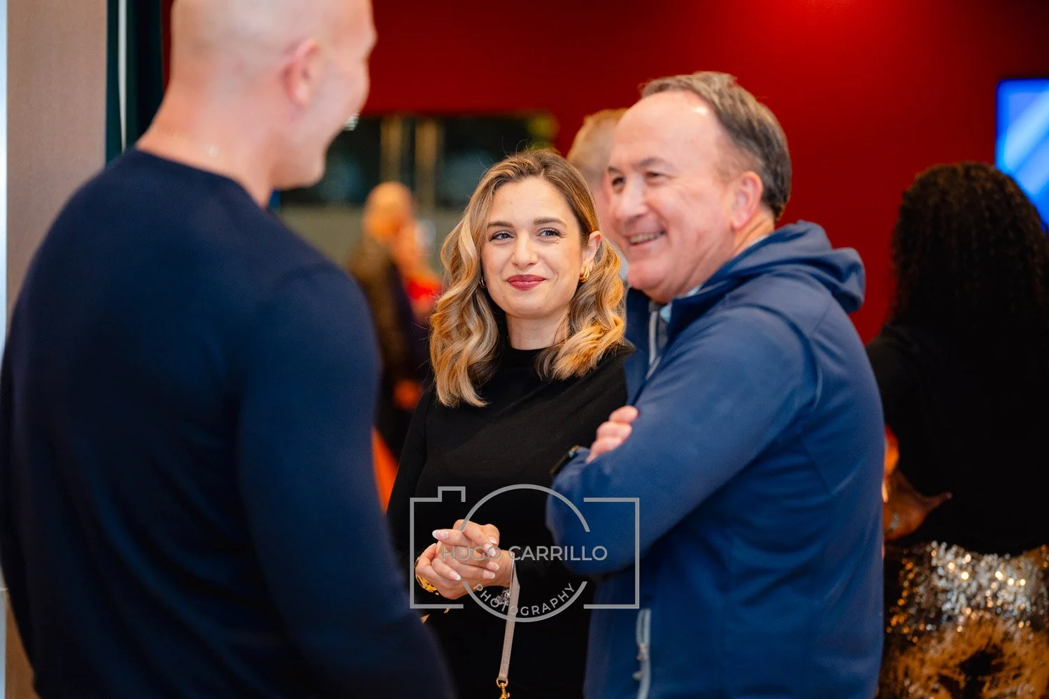 Three people talking at a social event, smiling and engaging in conversation, with a woman and a man in the foreground and another person in the background.