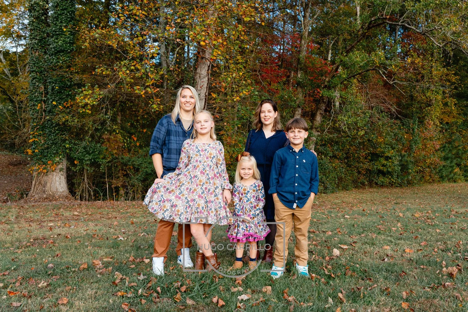 Family of six standing outdoors on a grassy area with fall foliage in the background, including two adult women, one teenage girl, one young girl, and two boys.