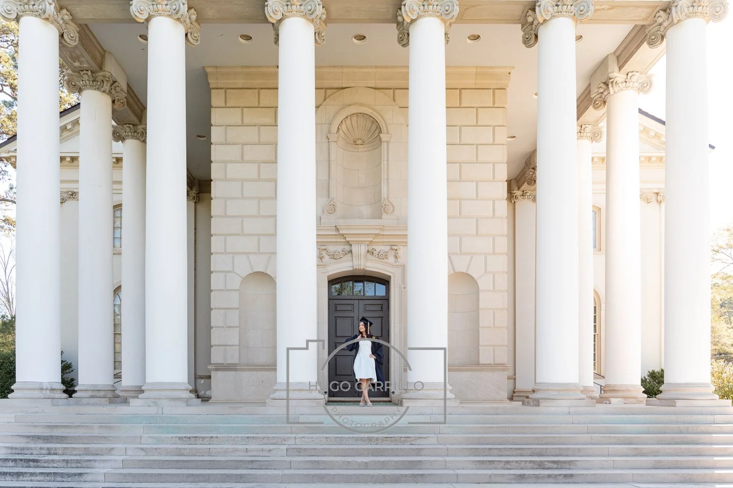 A woman in graduation attire standing on steps in front of a large, classical-style building with tall white columns.