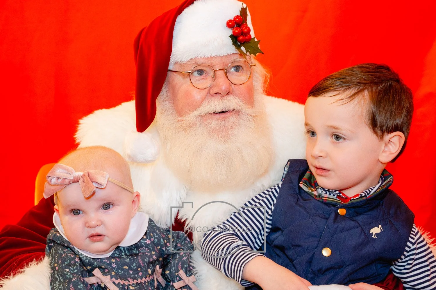 Santa Claus with a baby girl and a young boy sitting together in front of a red background.
