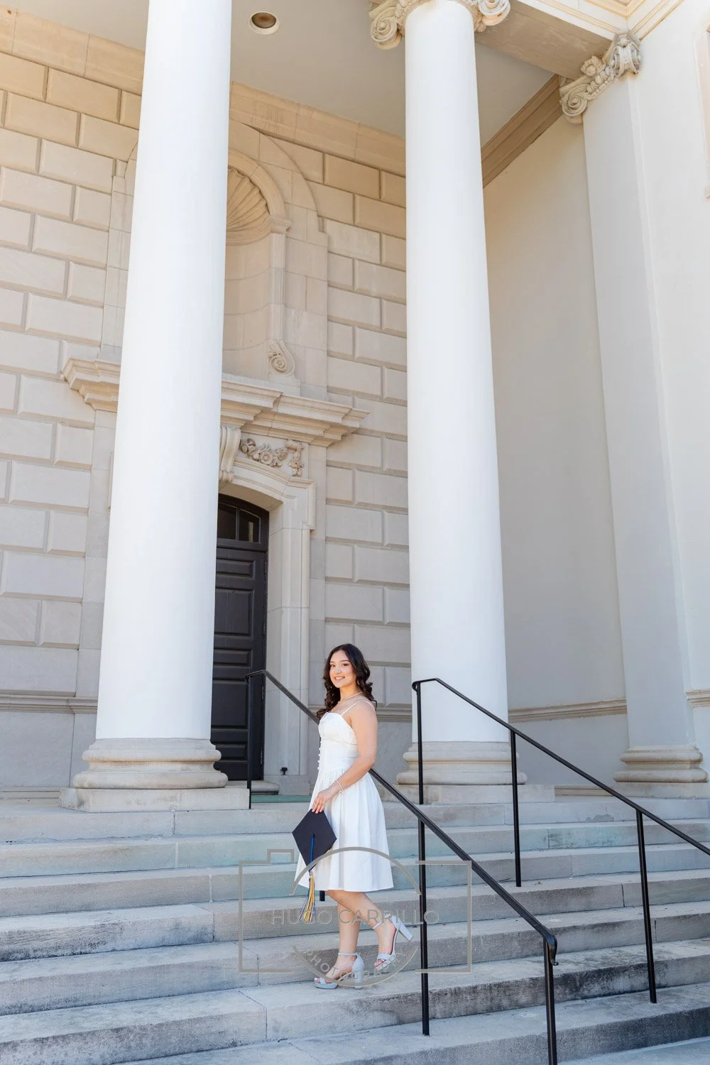 A young woman in a white dress and high heels holding a graduation cap standing on steps in front of a large building with tall white columns.