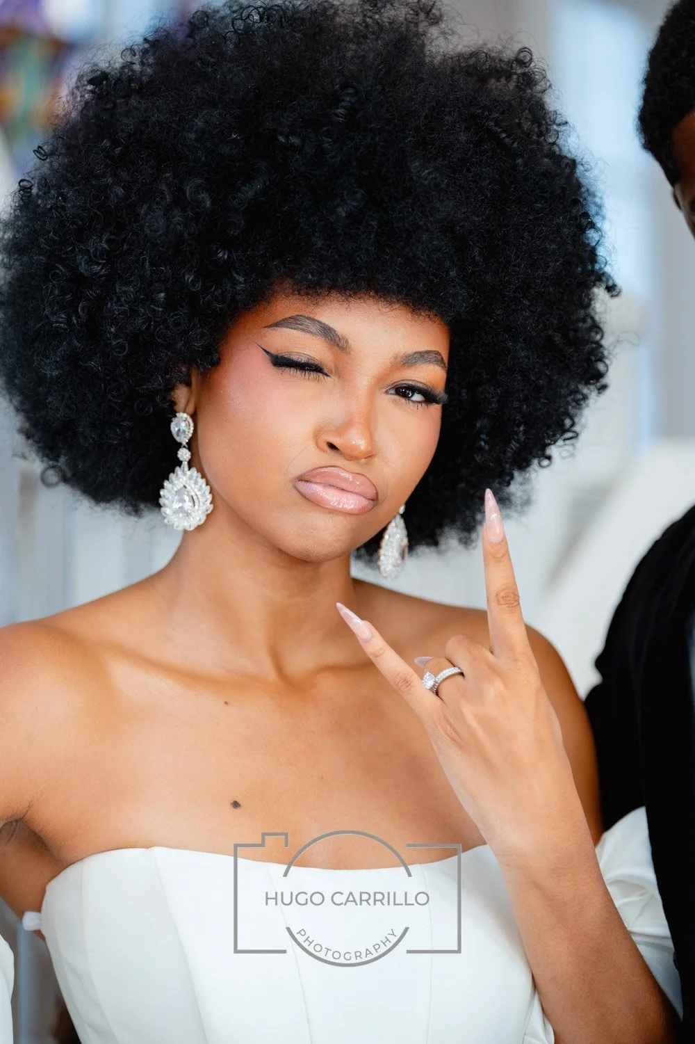 A woman with a large, curly black hairstyle is making a playful rock sign with her right hand, one eye closed and lips slightly pursed. She is wearing elegant earrings, rings, and a white off-the-shoulder top, with a confident expression.