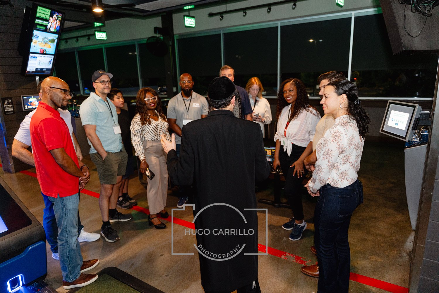 A group of diverse people gathered around a man in a black coat and hat, who appears to be speaking or giving a presentation in a modern indoor space with large windows and electronic screens.