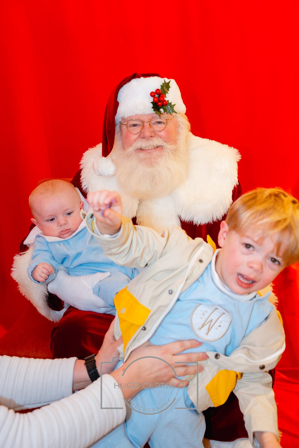 Santa Claus sitting with two young children against a red background, one child sitting on Santa's lap and the other being held in front, both children dressed casually.