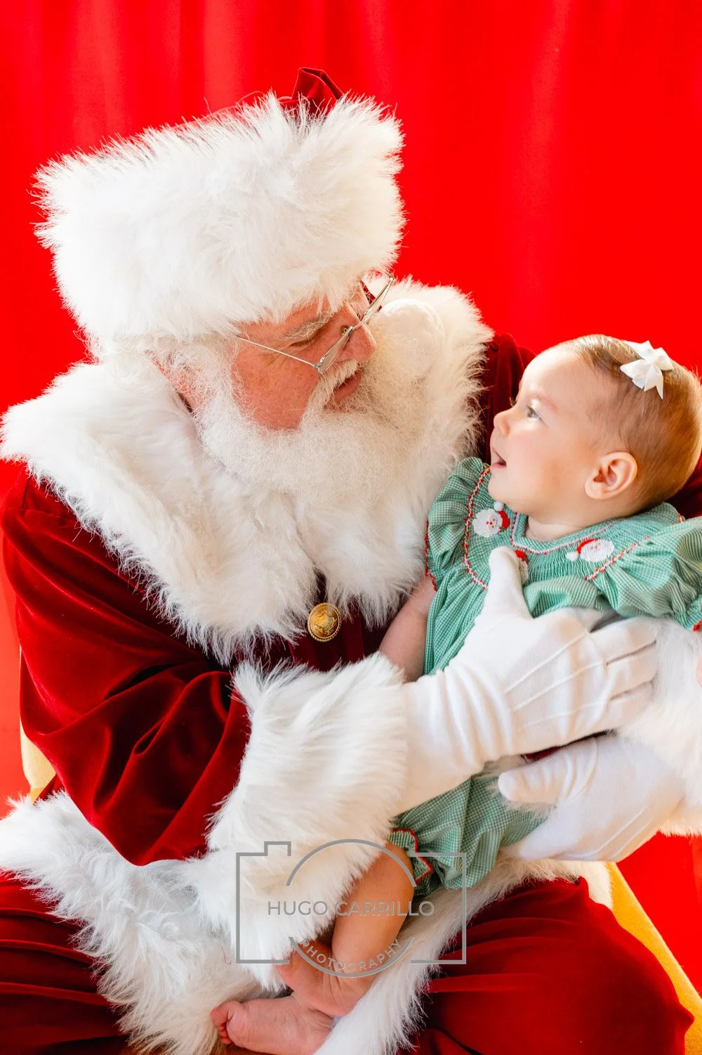 Santa Claus in a red suit with white fur trim holding a young girl with a white bow in her hair against a red background.