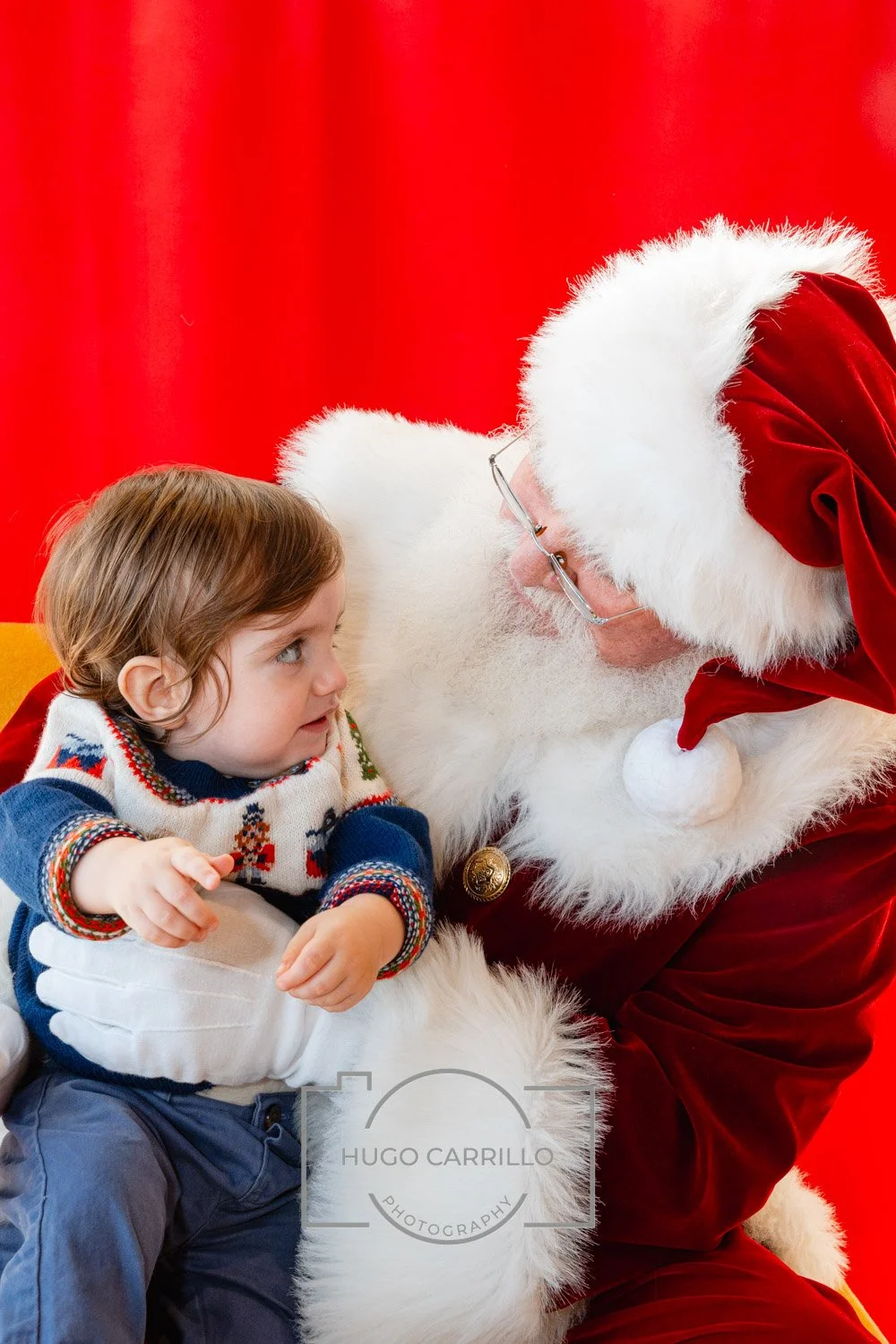 A young girl talking to Santa Claus, who is dressed in a red velvet suit with white fur trim, in front of a red background.