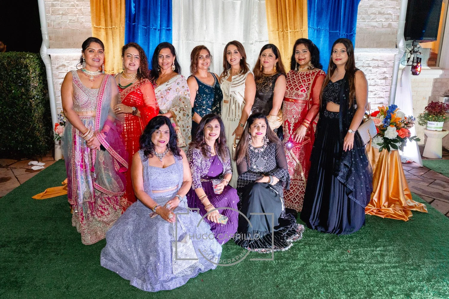 Group of women dressed in colorful traditional Indian attire at an outdoor celebration, standing on artificial grass in front of a decorated backdrop with blue, yellow, and white curtains, and a table with flowers.