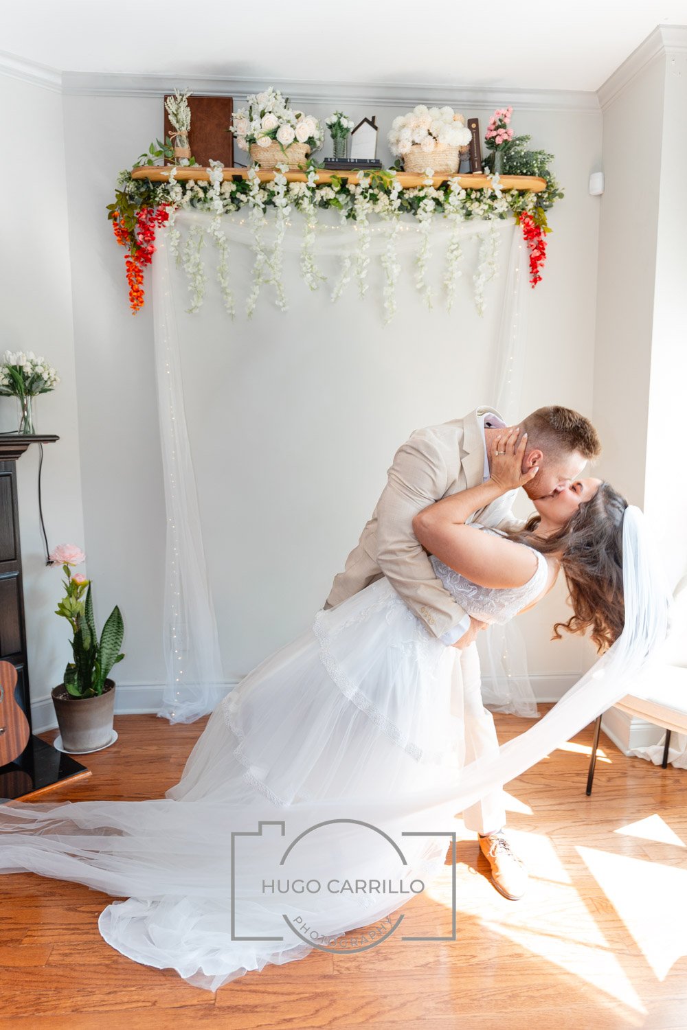 A bride and groom sharing a kiss in a room decorated with white and colorful flowers, with sunlight streaming in.