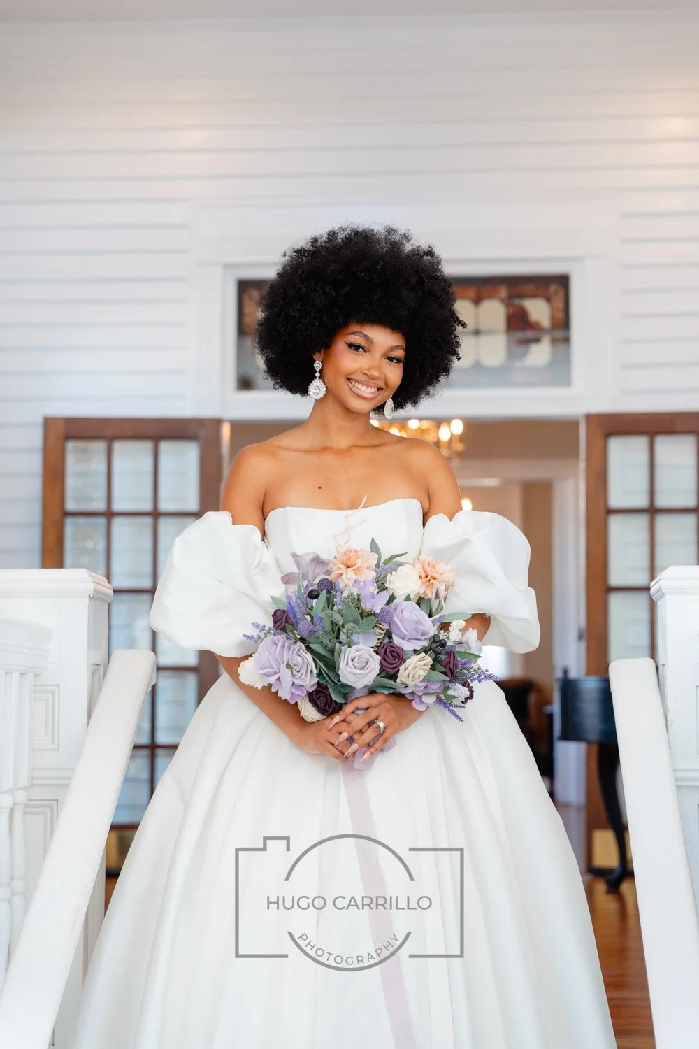 A woman in a white wedding gown holding a bouquet of purple, lavender, and blush flowers, smiling in a bright, indoor setting.