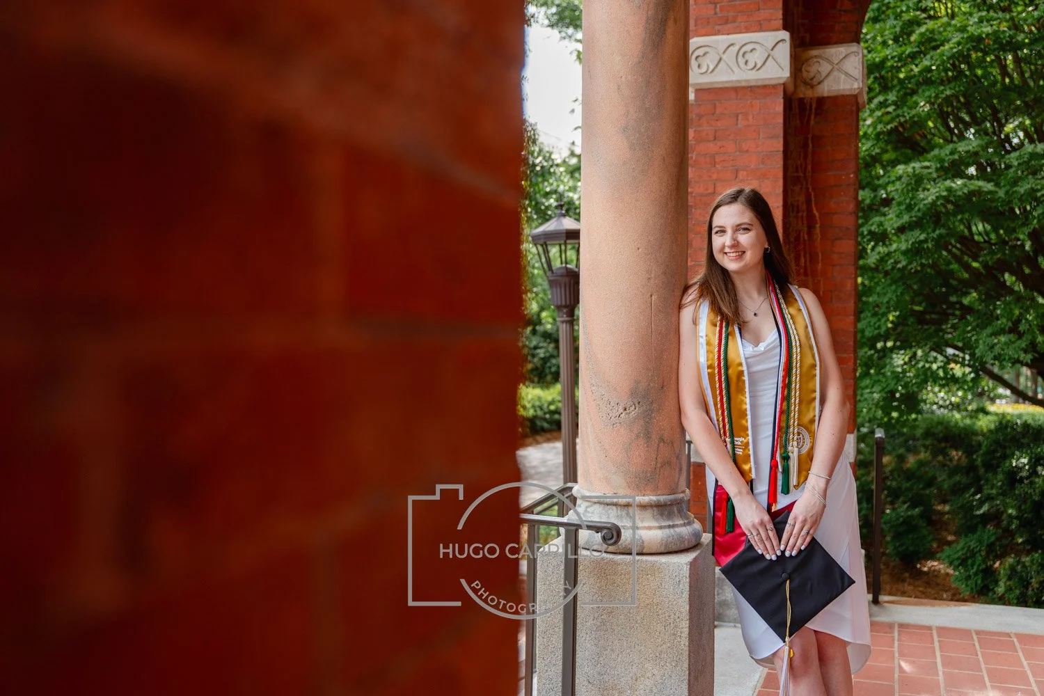 Young woman in graduation cap and gown, smiling and standing outdoors beside a brick column and greenery.