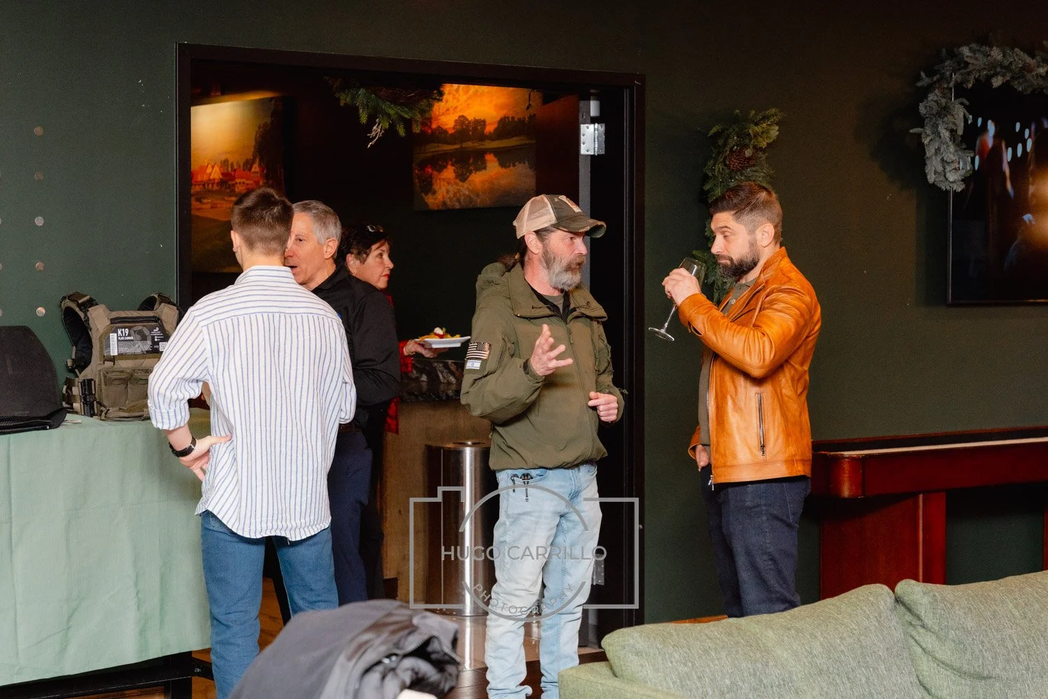 Group of five people socializing at an indoor gathering, some holding drinks, with a green wall, a window with a sunset view, and holiday decorations in the background.