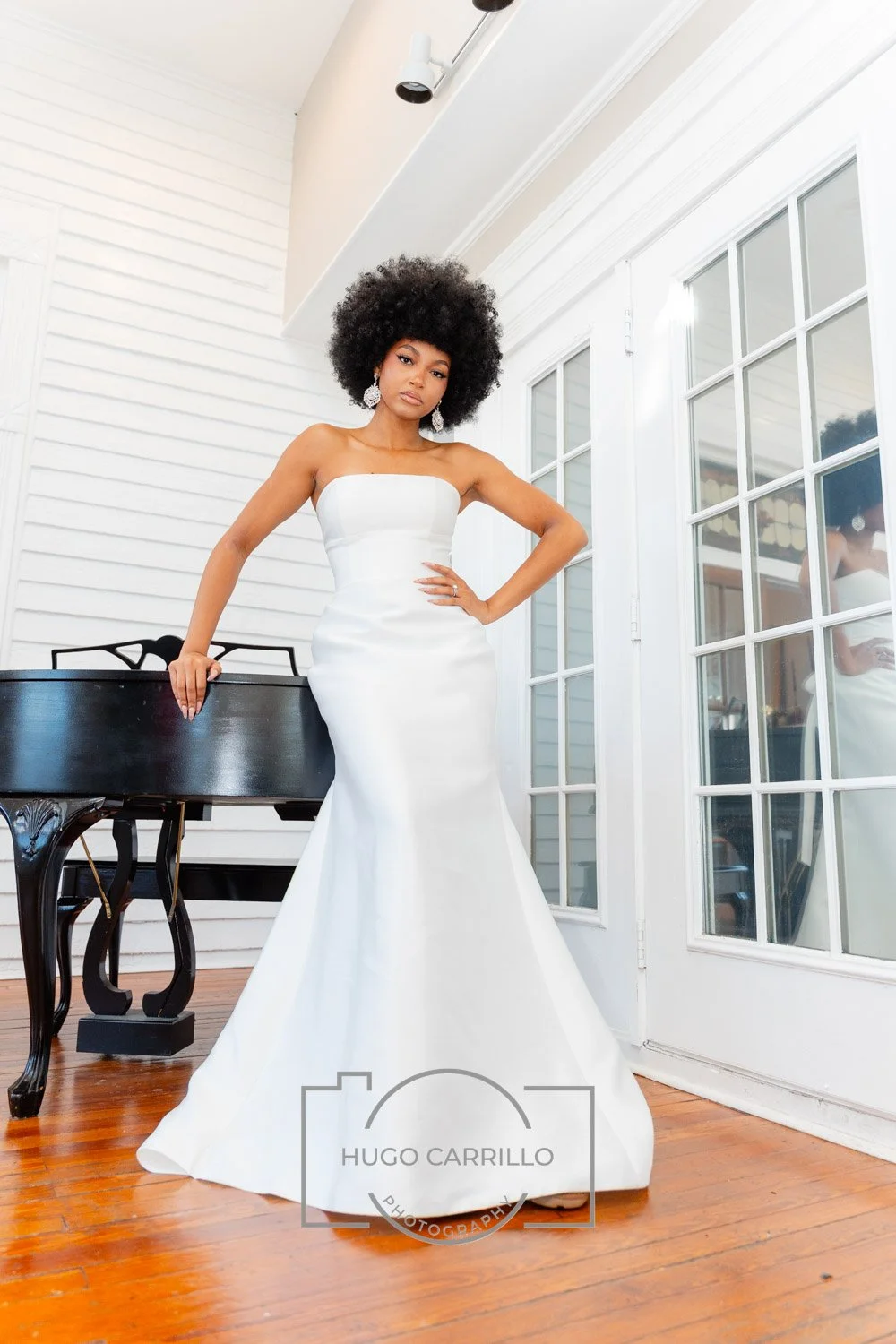 A woman in a strapless white wedding dress standing beside a piano on a wooden floor inside a room with white paneled walls and glass-paneled doors.