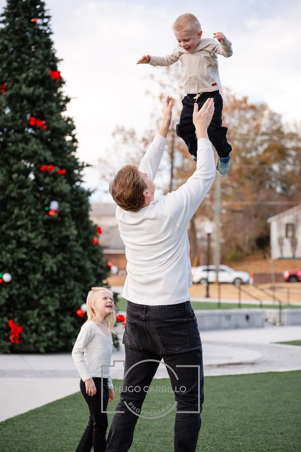 A man is lifting a young boy into the air while a young girl looks on happily. A decorated Christmas tree is visible in the background, indicating festive holiday cheer.
