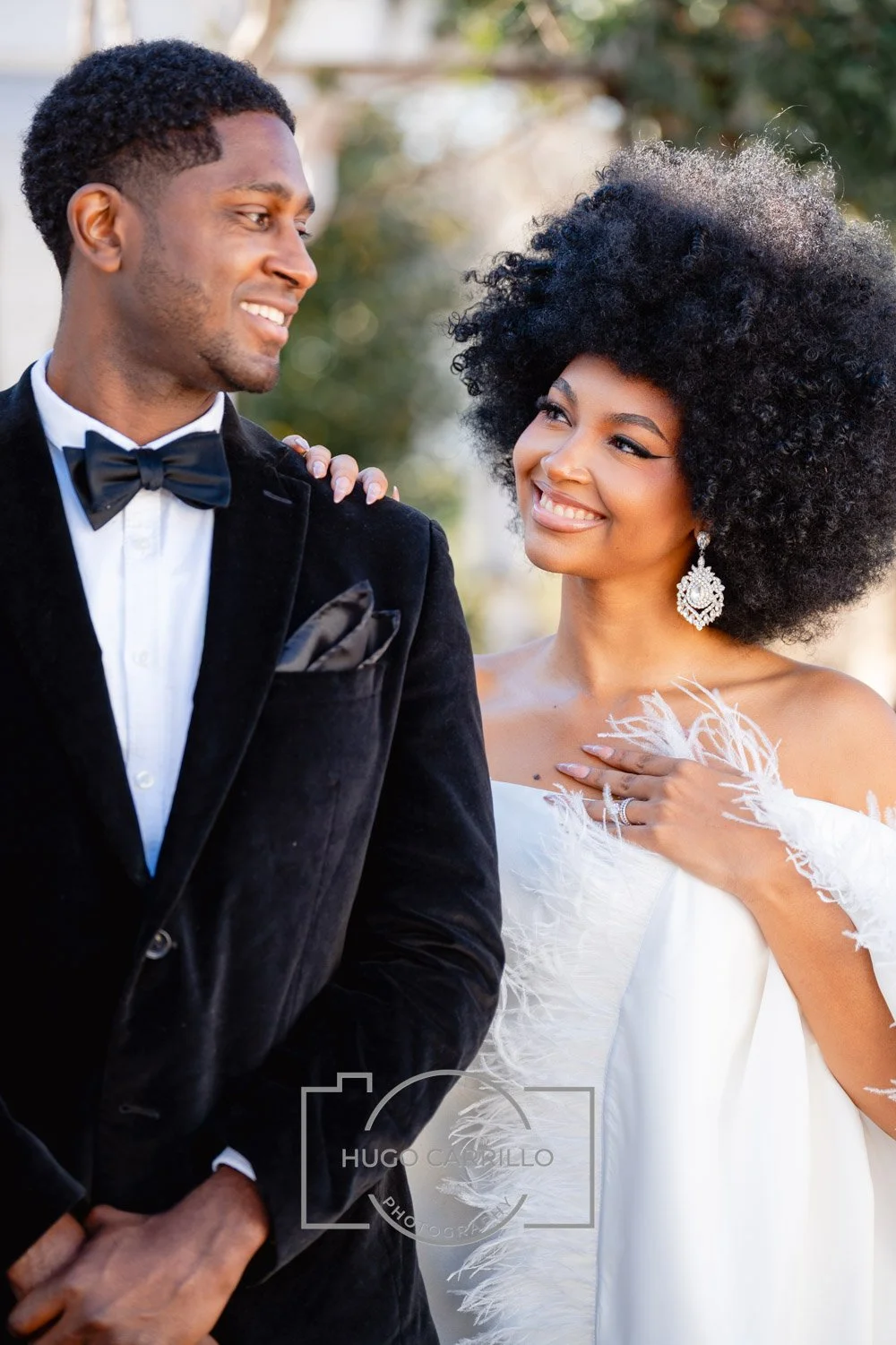 A joyful couple dressed in wedding attire, outdoors, with the woman in a white dress with feather details and the man in a tuxedo with a bow tie, sharing a tender moment.