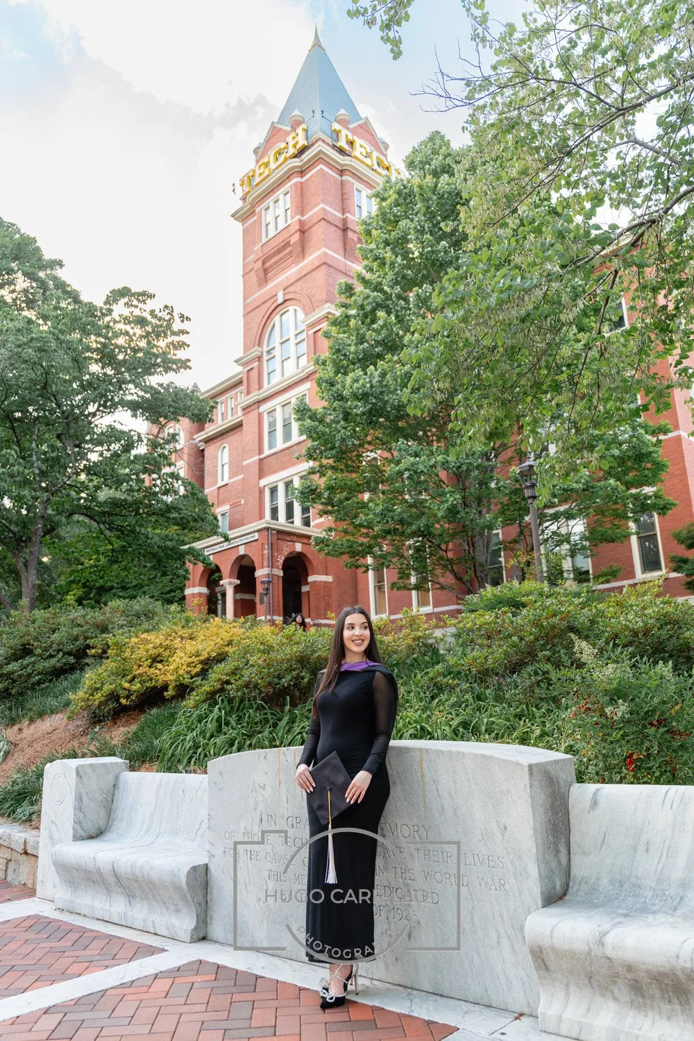 A young woman in a graduation gown standing on a concrete bench in front of a historic red-brick building with a clock tower, surrounded by trees and greenery.