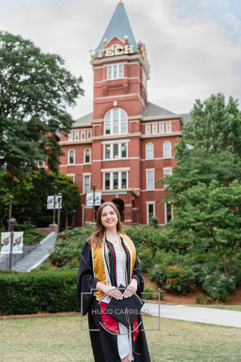 A young woman in a graduation gown and cap standing outdoors in front of a red brick university building, smiling.