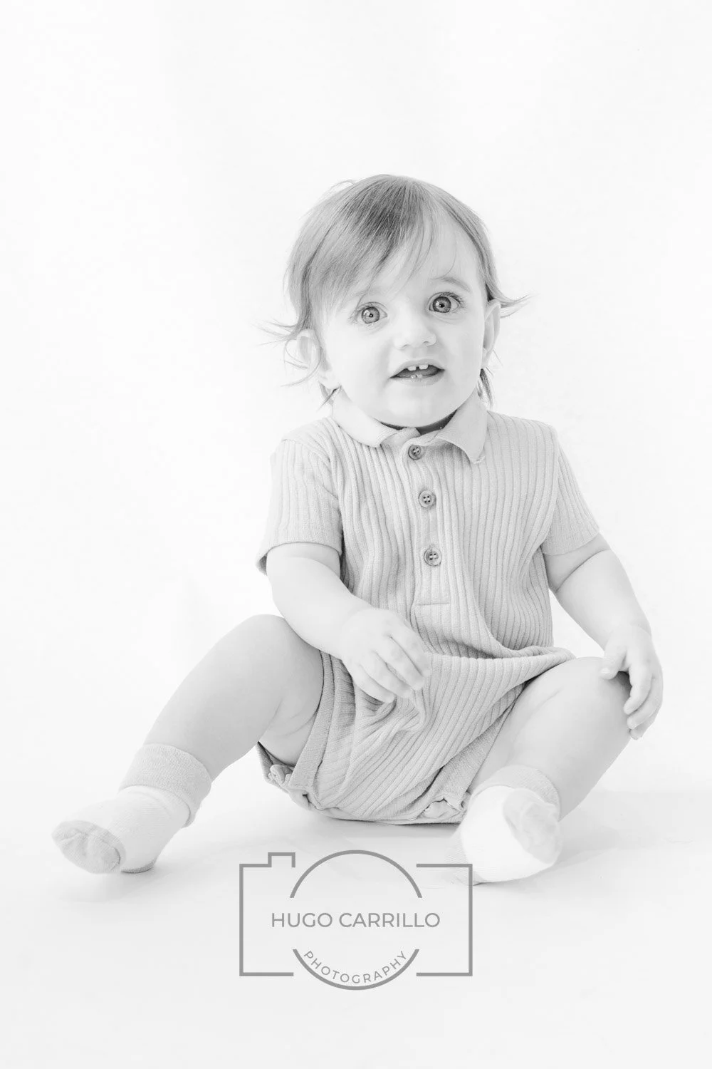 Black and white photo of a smiling toddler sitting on the floor with legs apart, wearing a striped polo shirt, shorts, and socks, in a studio setting with a plain background.