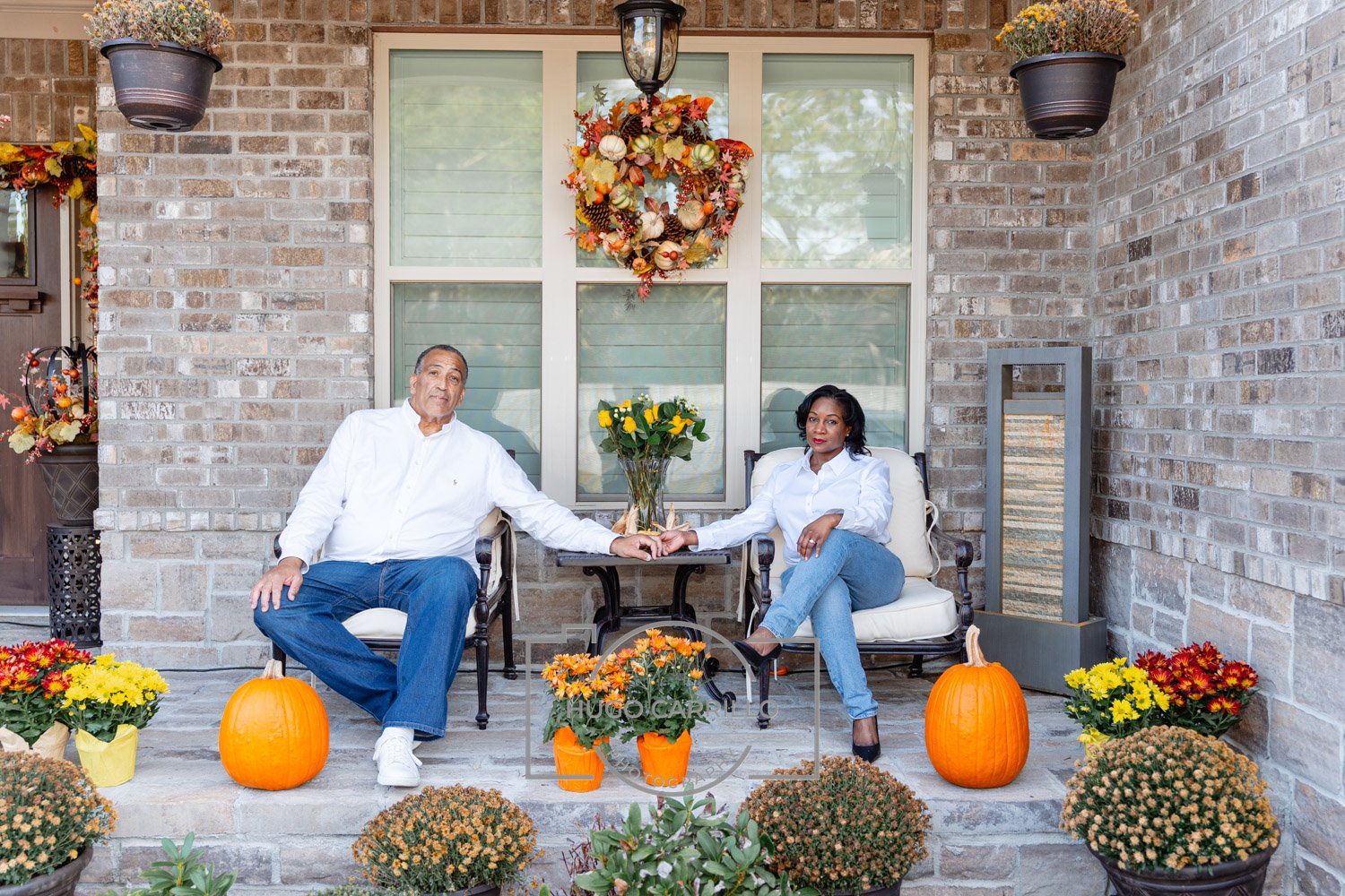 A man and woman sitting on a porch surrounded by pumpkins, flowers, and fall decorations, holding hands across a small table with a flower vase, during autumn.