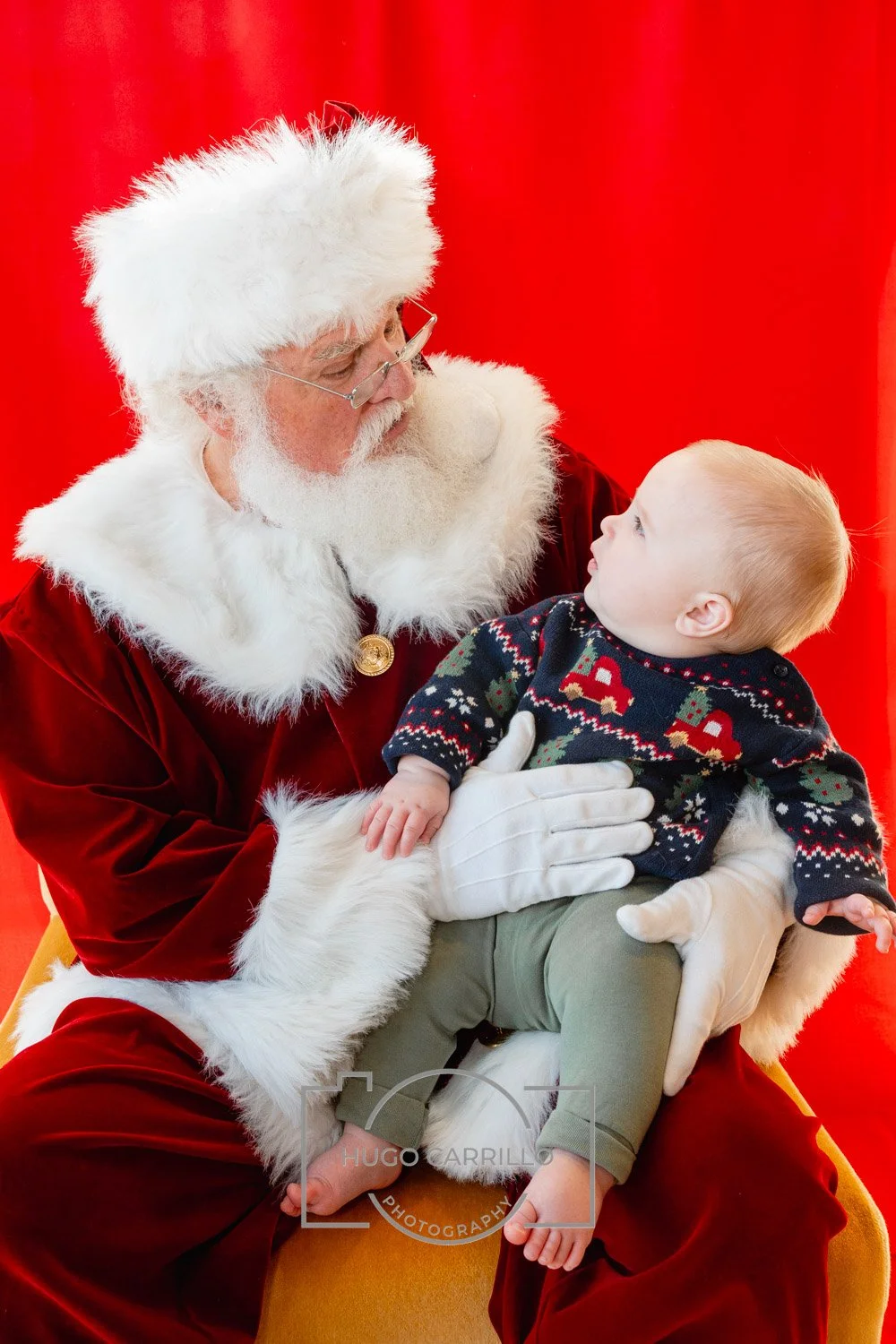 Santa Claus, with white hair and beard, wearing glasses, a red suit with white fur trim, holding a young child in holiday sweater, in front of red background.