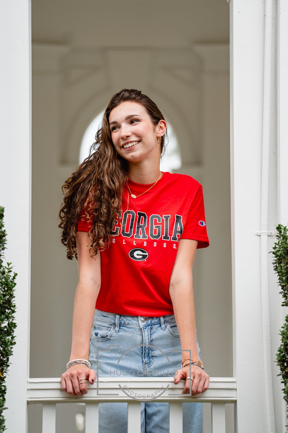 A young woman with long, curly brown hair smiling and leaning on a white railing, wearing a red Georgia Bulldogs T-shirt and light blue jeans.