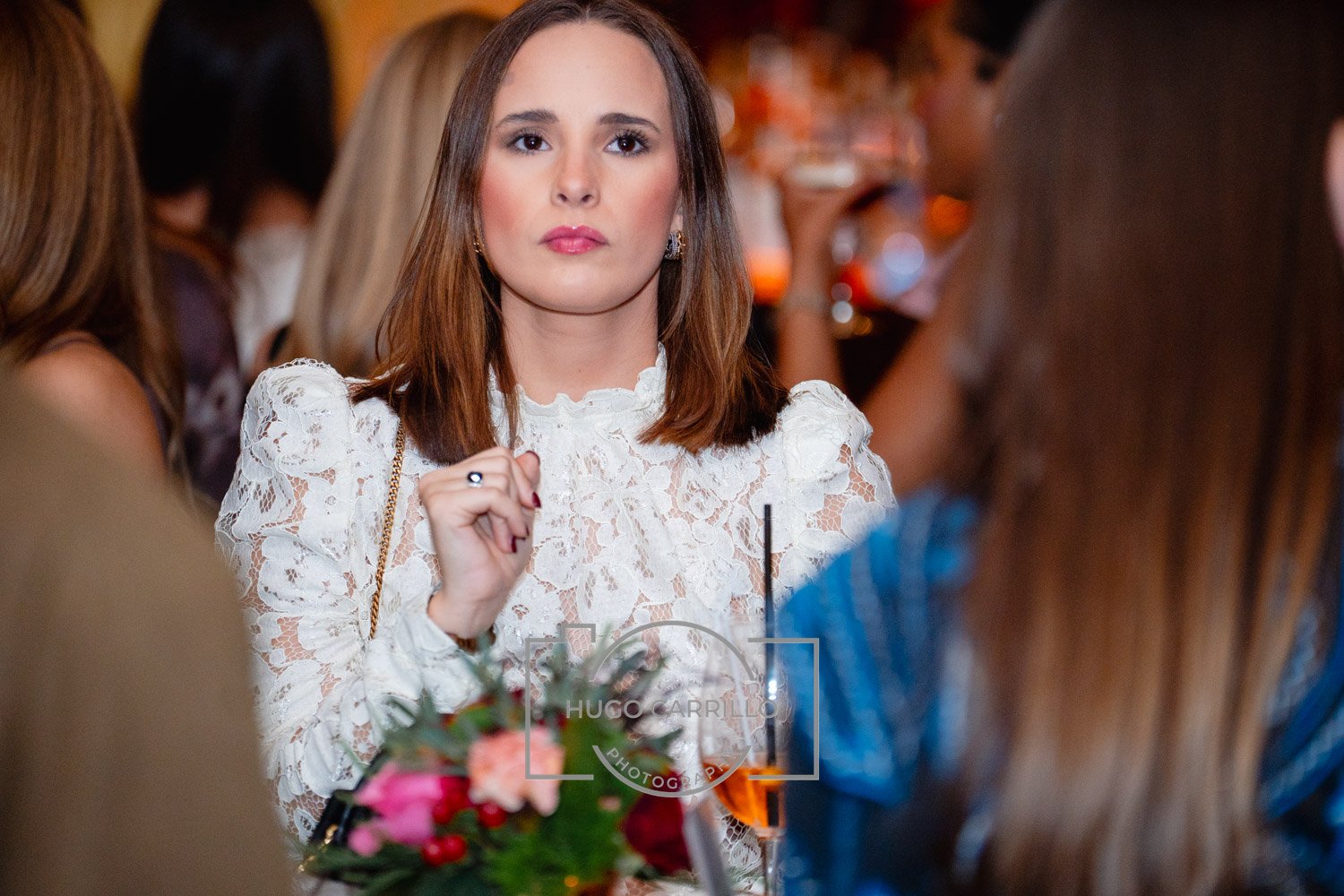 A woman with shoulder-length brown hair and a serious expression, wearing a white lace blouse, is sitting at a table with a floral centerpiece and a drink with a straw, in a crowded indoor setting.